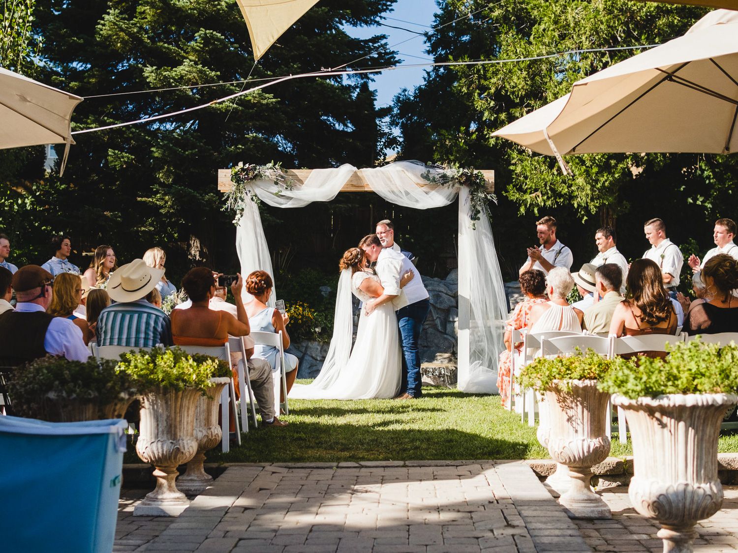 Outdoor wedding ceremony with couple kissing under floral arch, guests seated on garden benches, surrounded by lush greenery.