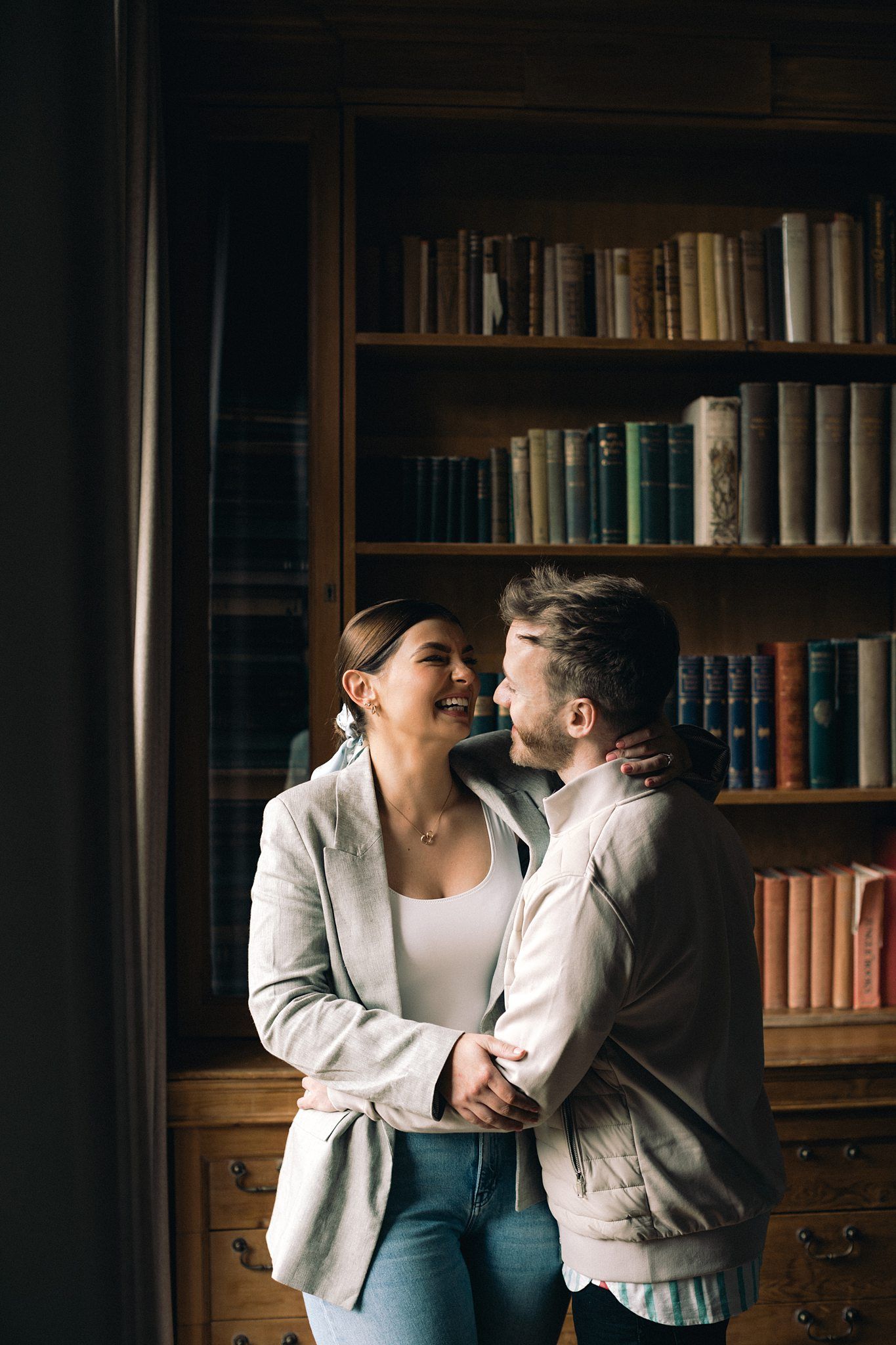 Couple embracing and smiling in a cozy library with wooden bookshelves filled with books in the background.