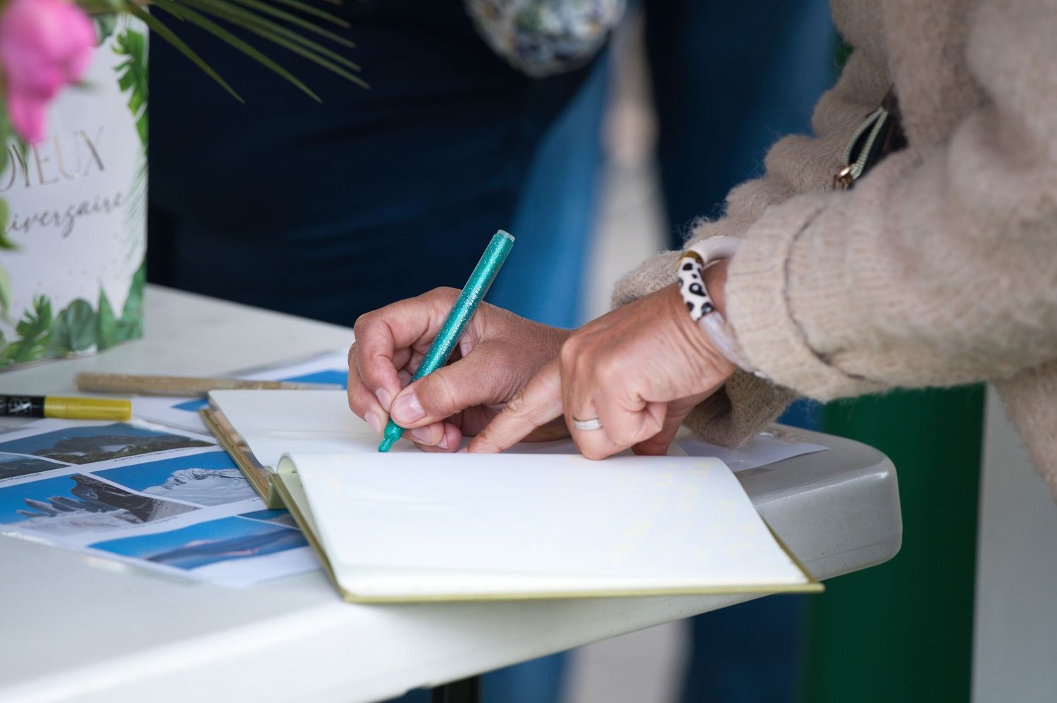 Close-up of hands writing in an open notebook with a teal pen, wearing a bracelet, at a white table.