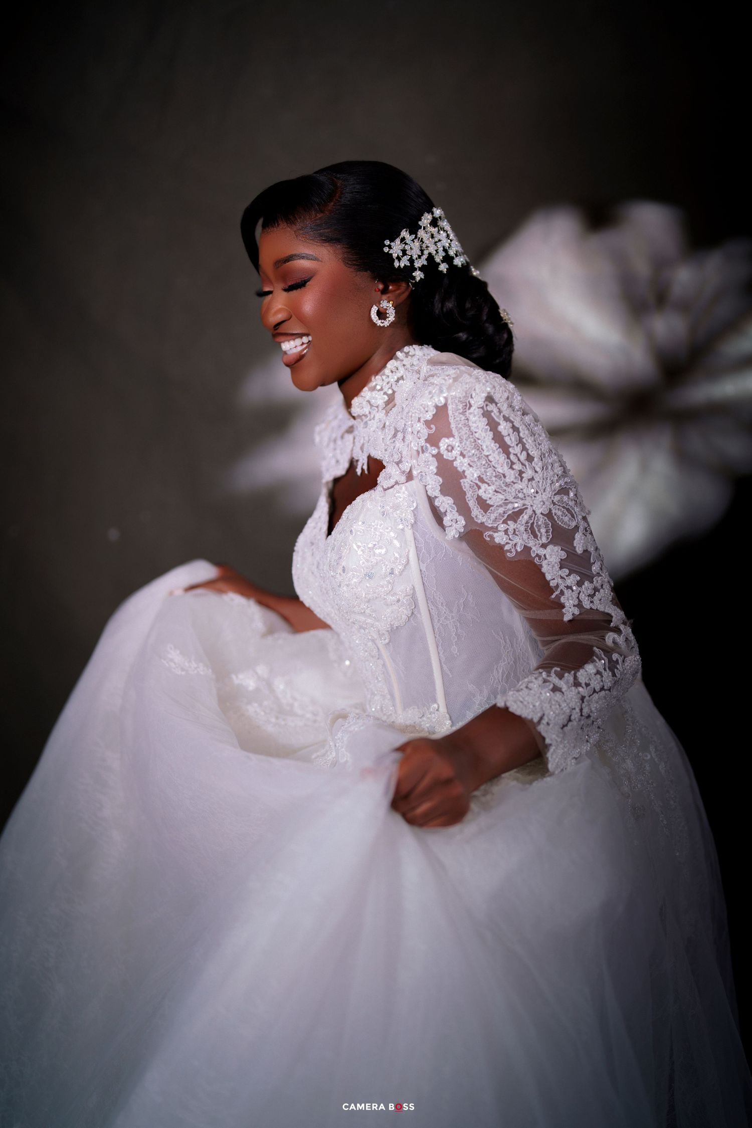 Smiling bride in white lace ball gown with sheer sleeves and crystal hair accessory poses joyfully.