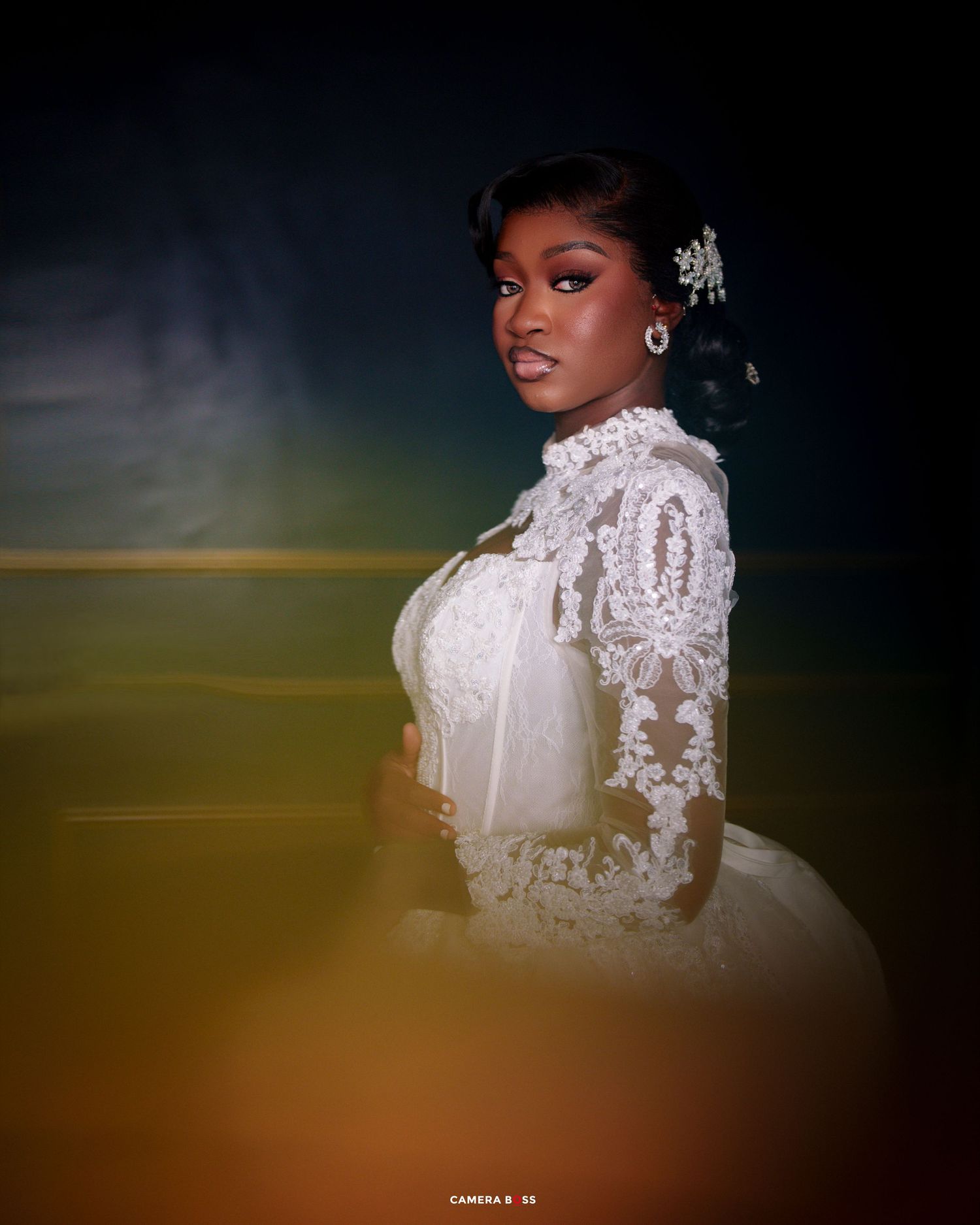 Elegant bride in white lace long-sleeve wedding gown with floral hair piece poses gracefully indoors.