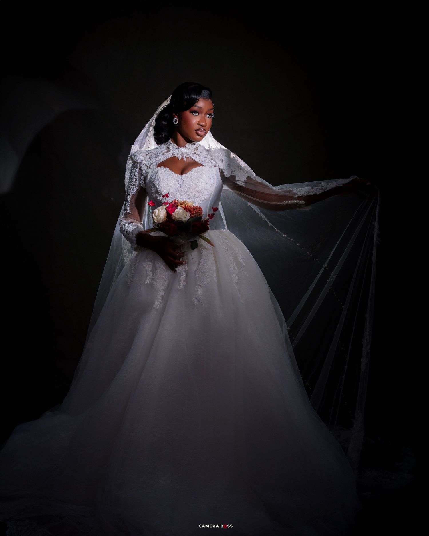 Bride in white lace ball gown wedding dress with veil holding colorful bouquet against dark background.