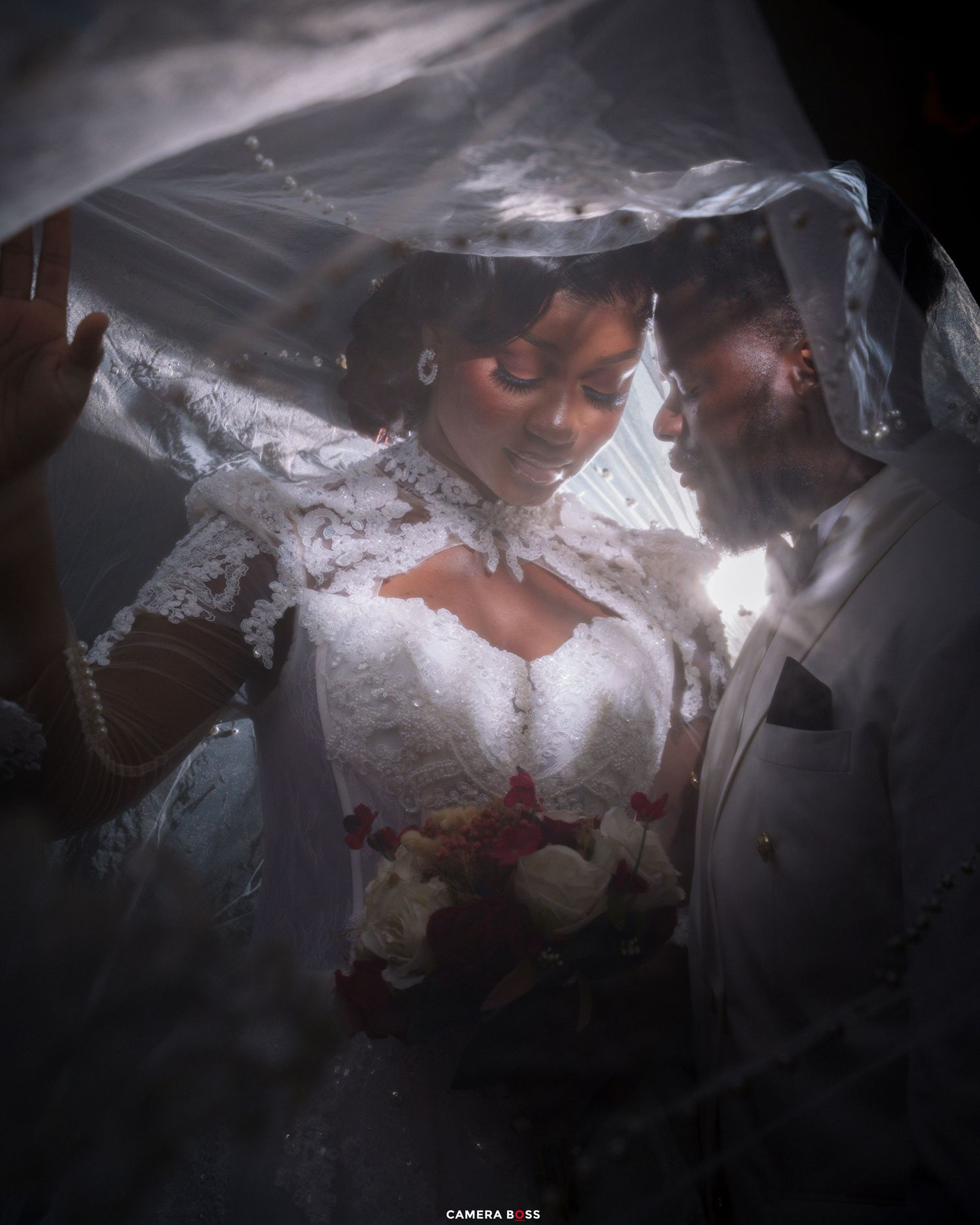 Bride and groom share an intimate moment under a white wedding veil, bride holding a bouquet of red and white flowers.
