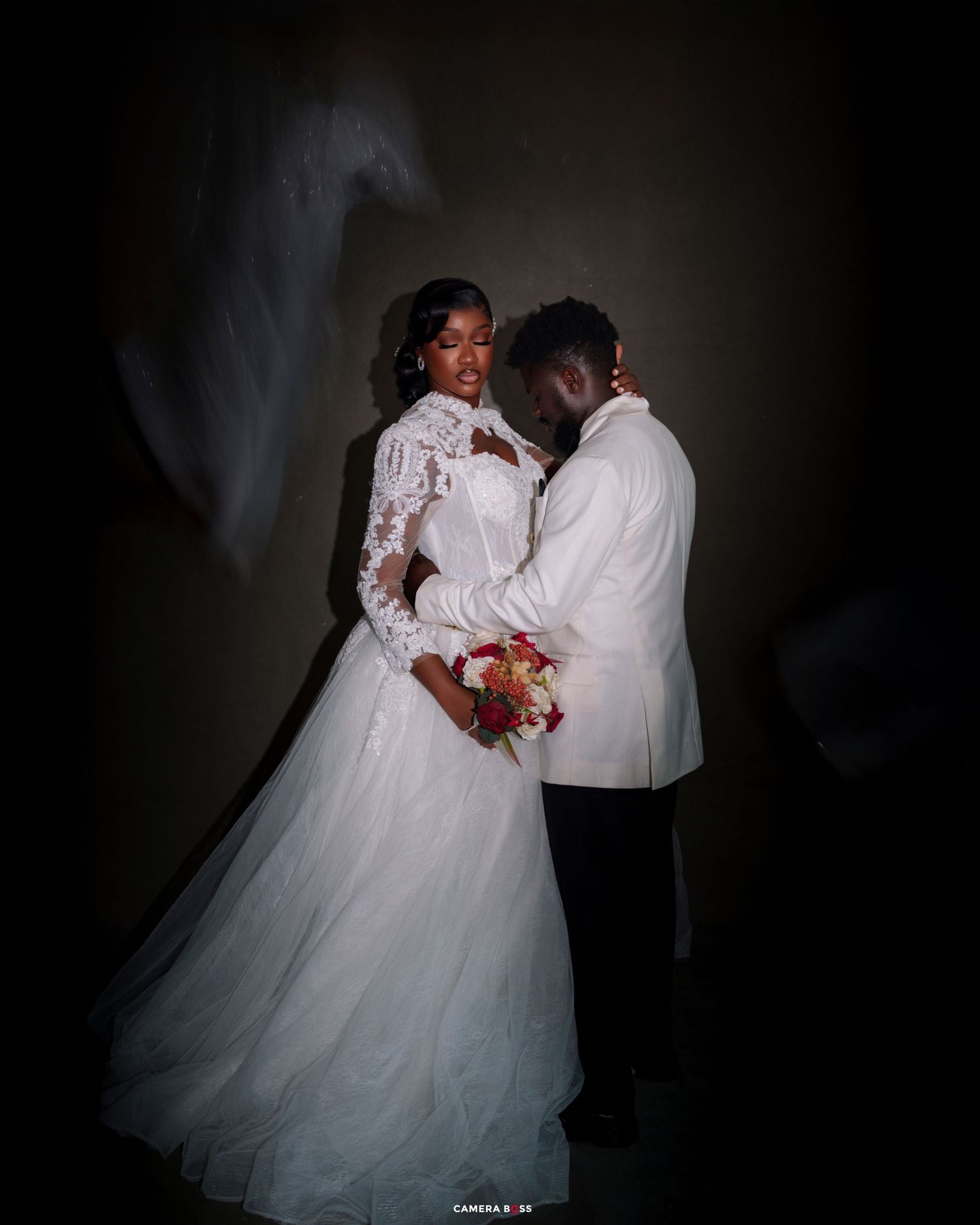 Elegant bride and groom in white wedding attire pose romantically against a dark background holding a bouquet.