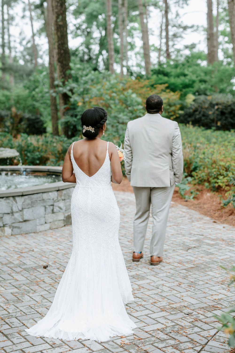 Bride in white backless gown and groom in tan suit walk hand in hand on brick path surrounded by lush greenery.