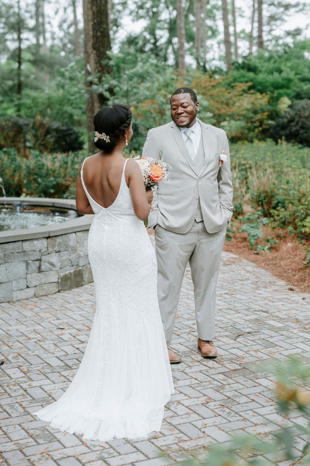 A smiling groom in a gray suit reacts joyfully as his bride in a white backless gown approaches during their first look.