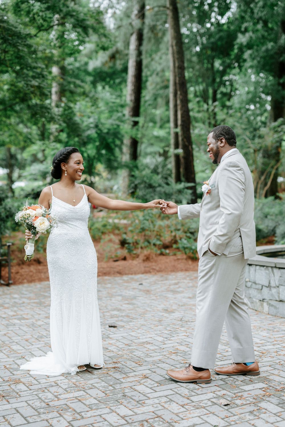 A bride and groom hold hands and smile at each other outdoors, surrounded by lush green trees on their wedding day.