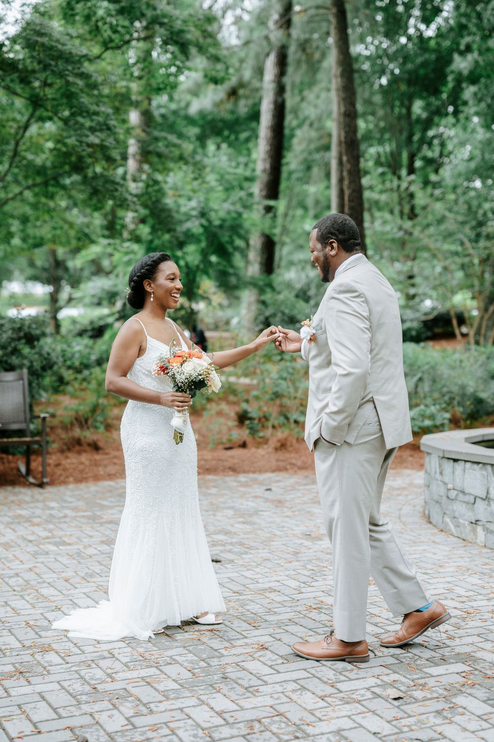 A joyful bride in a white gown holds a bouquet while sharing a first look moment with her groom in a lush garden.