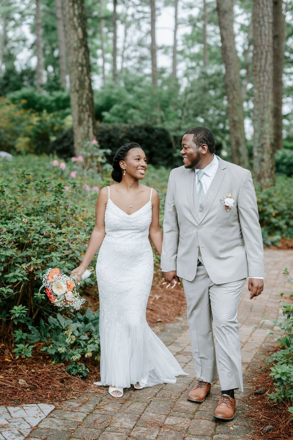 A smiling bride in a white gown and groom in a gray suit walk hand in hand along a brick path in a lush garden.
