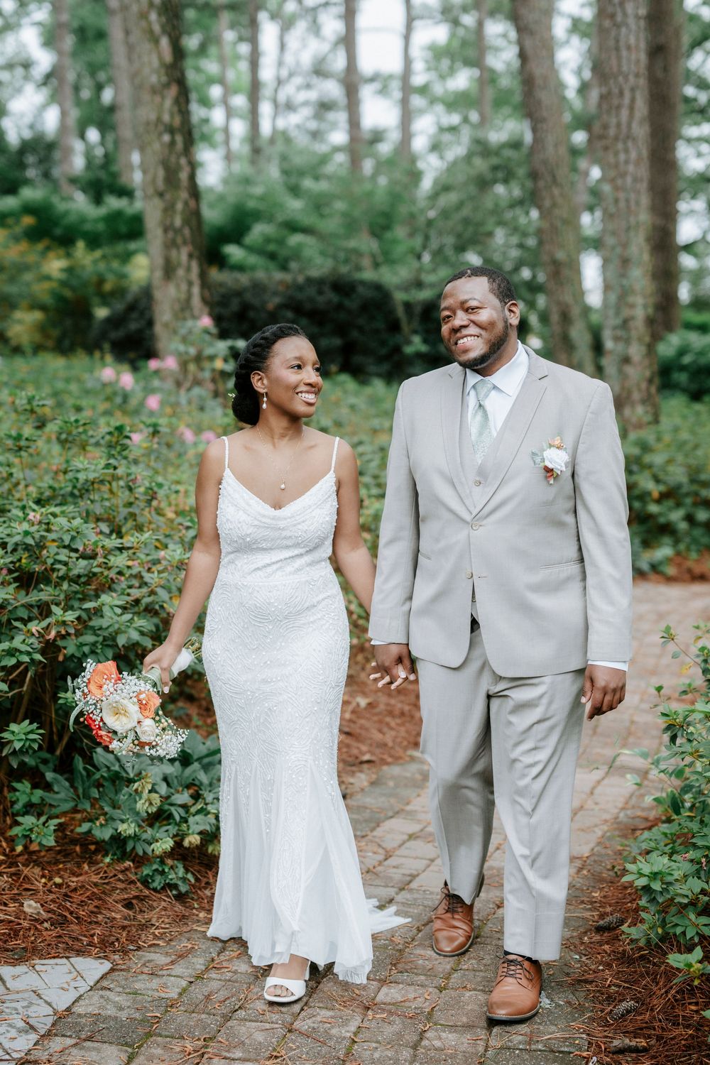 A smiling bride and groom walk hand-in-hand on a brick path surrounded by lush greenery on their wedding day.