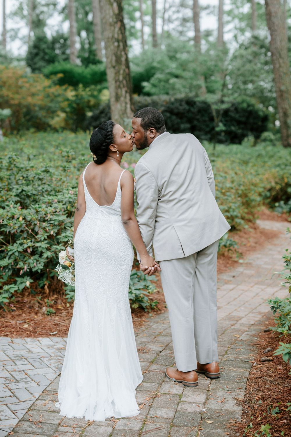 A newlywed couple shares a kiss on a brick path surrounded by lush greenery, bride in white gown holding bouquet.