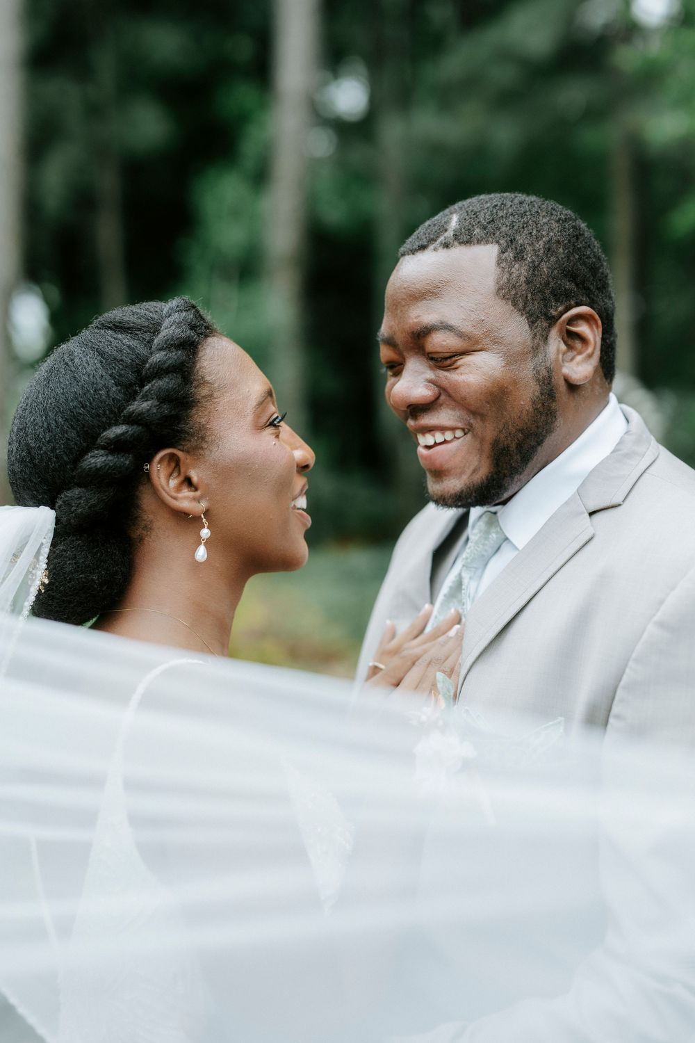 A joyful bride and groom share a loving gaze on their wedding day, with a flowing veil and lush green trees behind them.