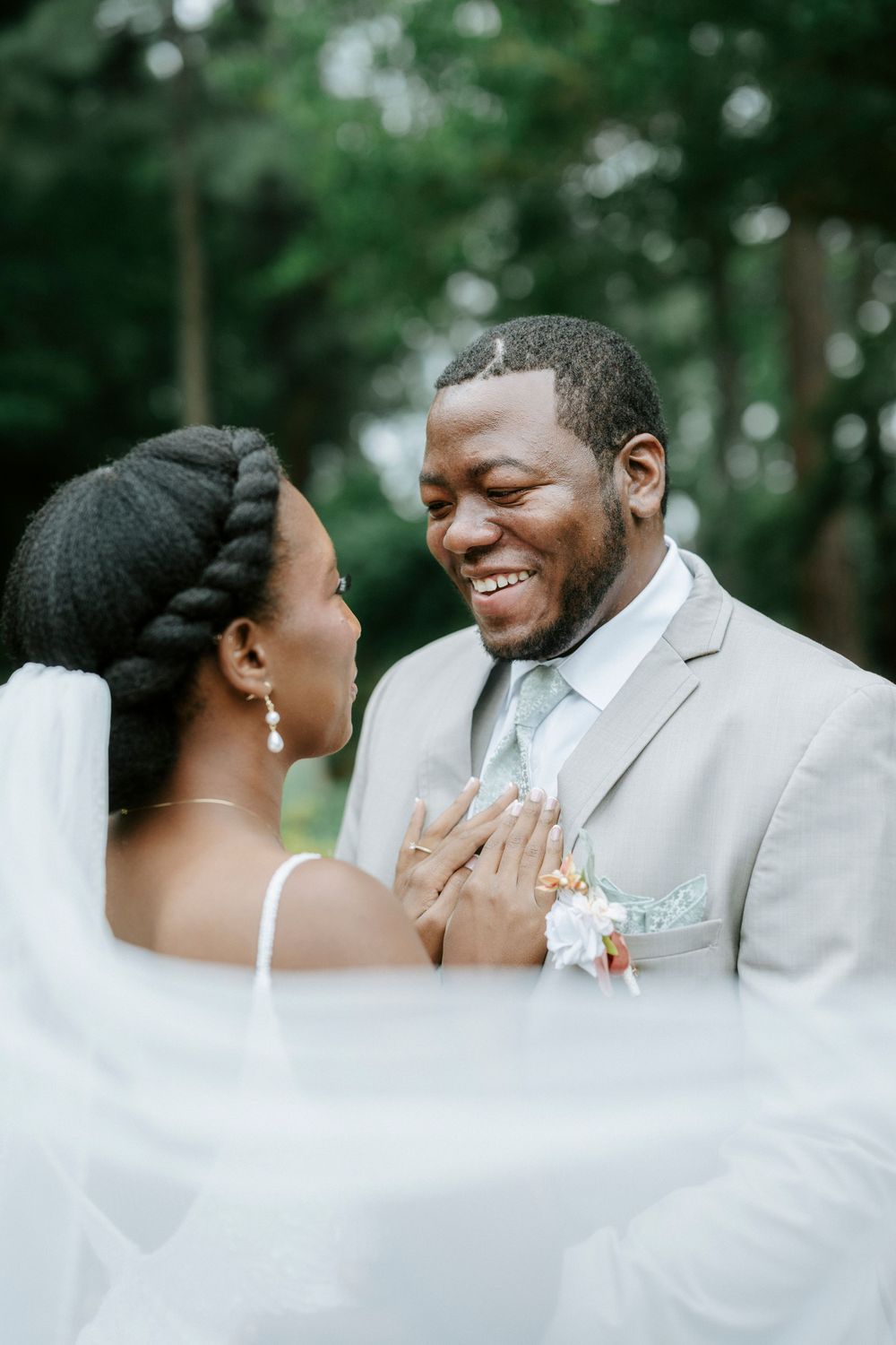 Joyful Black groom in gray suit laughs as bride in white veil and braided updo holds his hands outdoors.