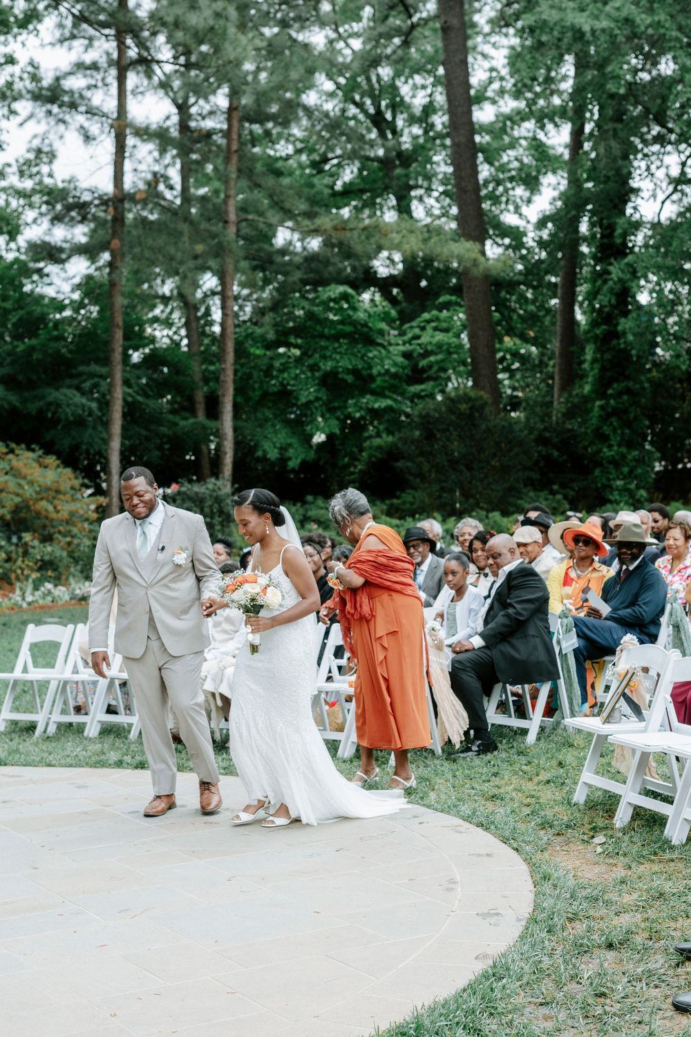 Bride and groom walk down the aisle at an outdoor wedding ceremony surrounded by lush green trees and seated guests.