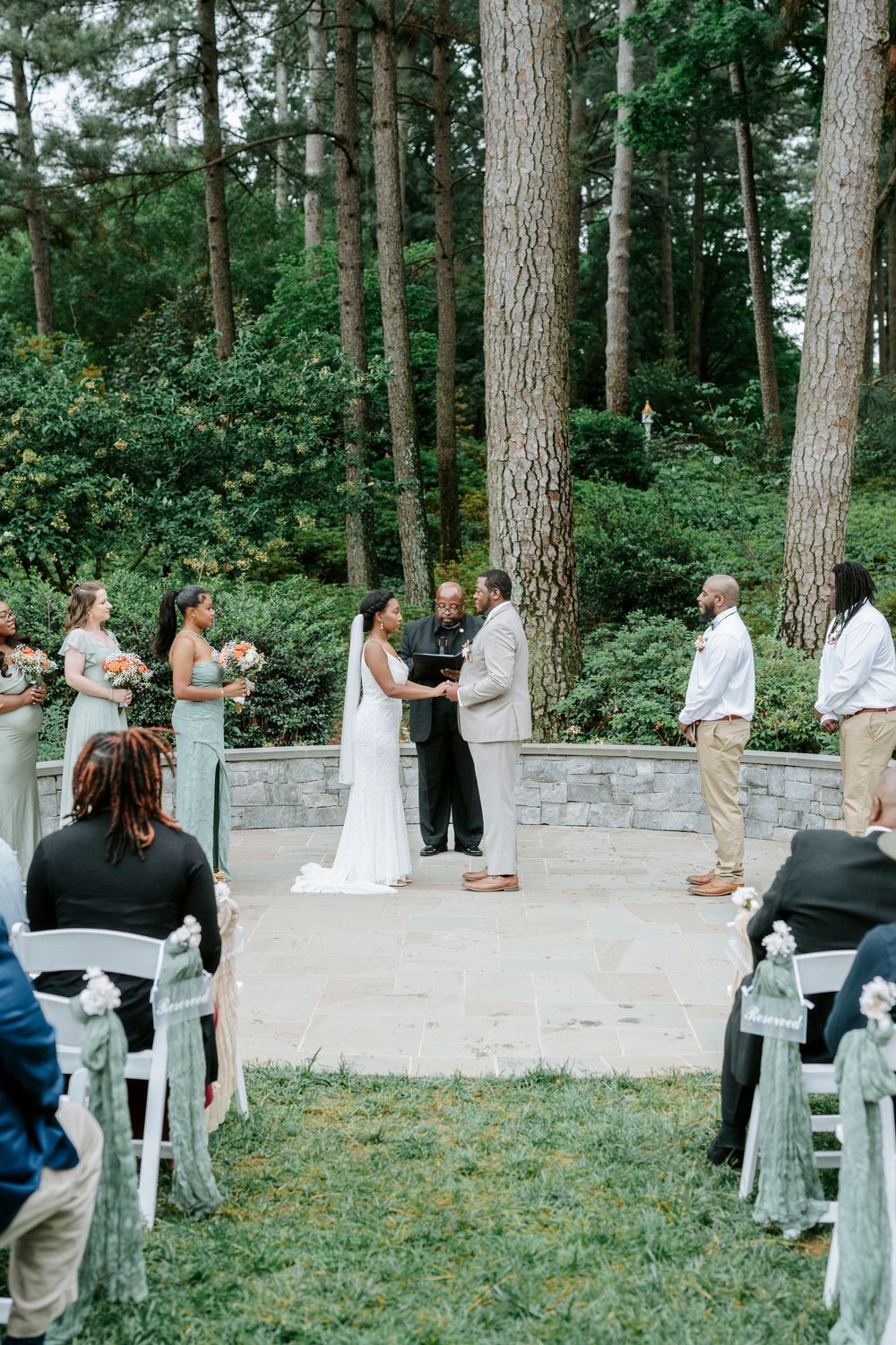 Outdoor wedding ceremony in a forest garden with bride, groom, officiant, bridesmaids, and seated guests on white chairs.