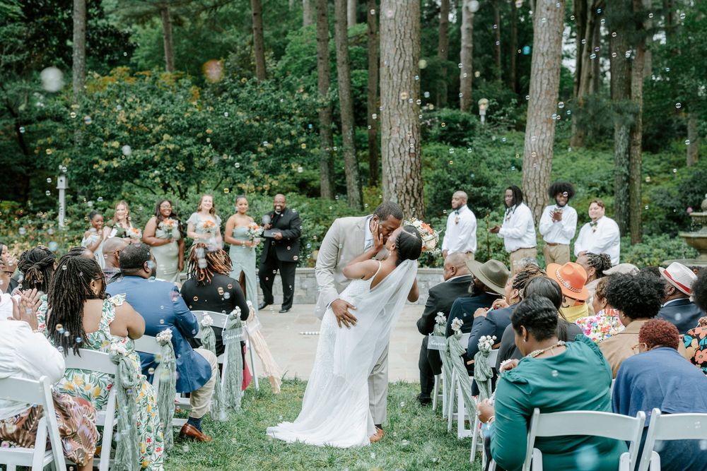 Bride and groom share their first kiss during an outdoor wedding ceremony surrounded by guests and tall trees.