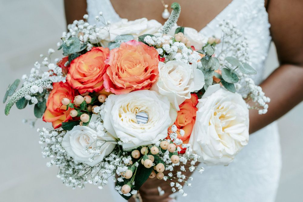 Bride holding a wedding bouquet with coral and white roses, baby's breath, and greenery, with gold rings nestled inside.