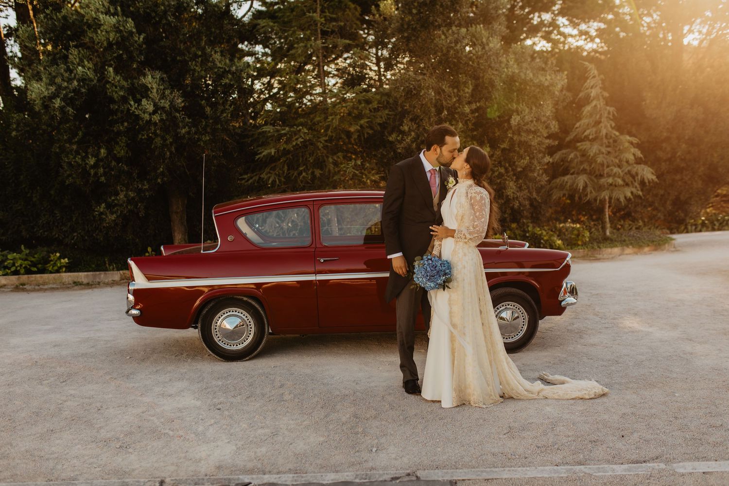 Bride and groom kissing beside a vintage red classic car at golden hour with lush green trees in background.