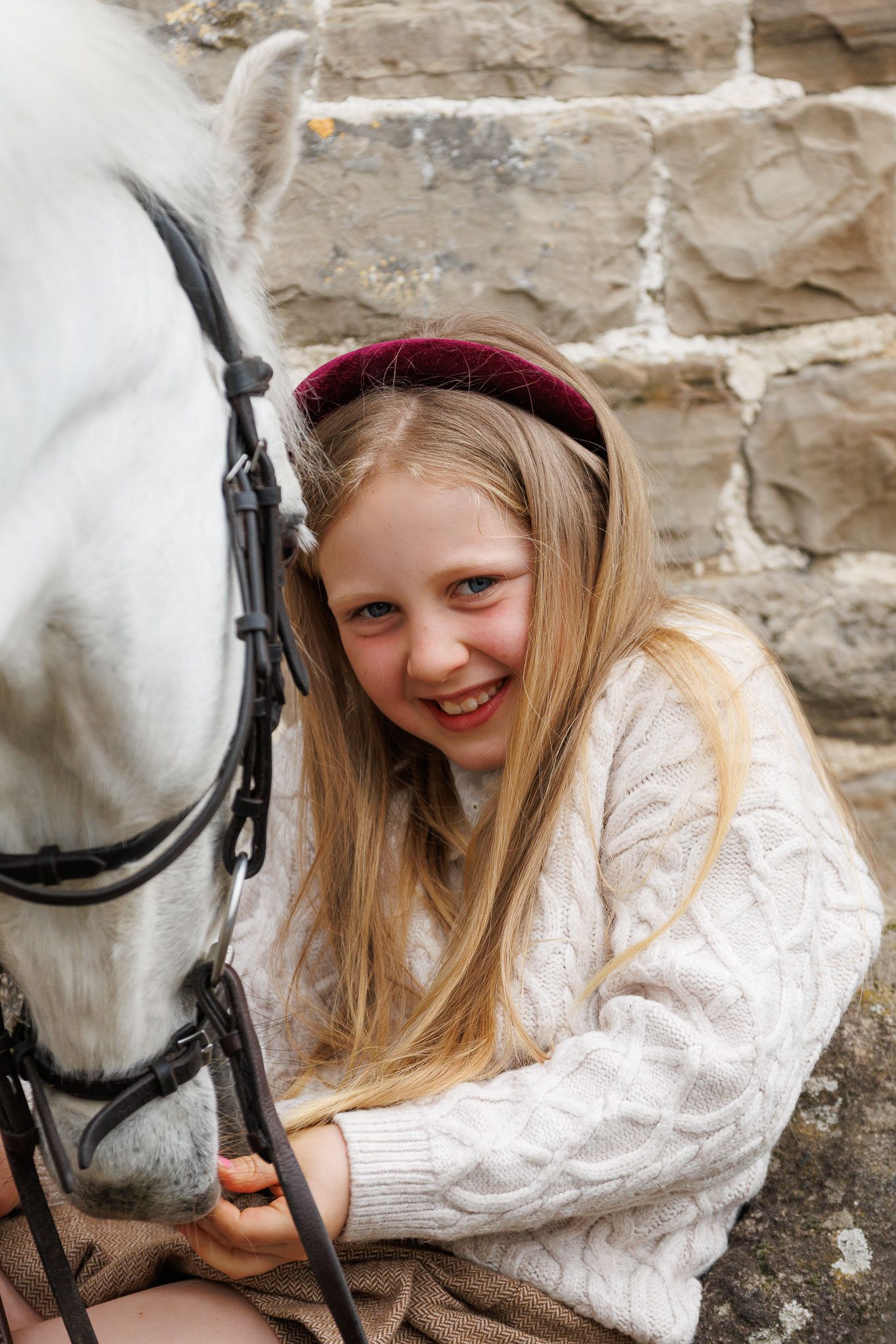 Smiling blonde girl in white cable knit sweater with red headband sitting beside a white horse near a stone wall.