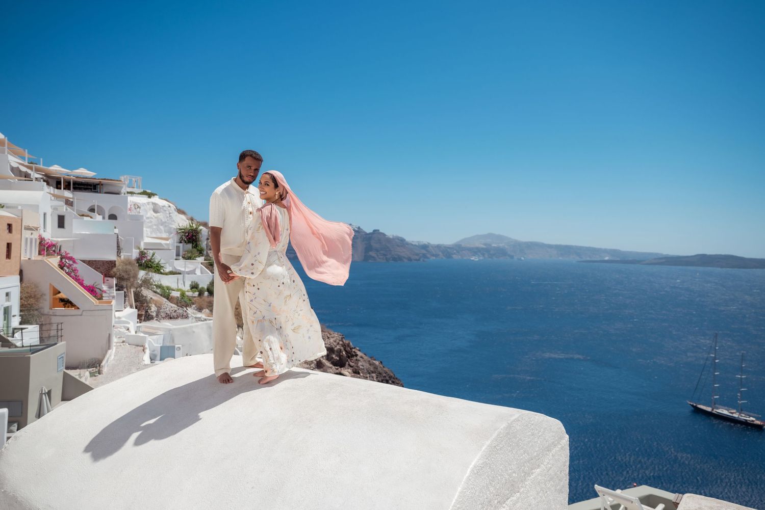 Couple poses on white rooftop in Santorini Greece with pink veil flowing in wind overlooking caldera and blue sea.