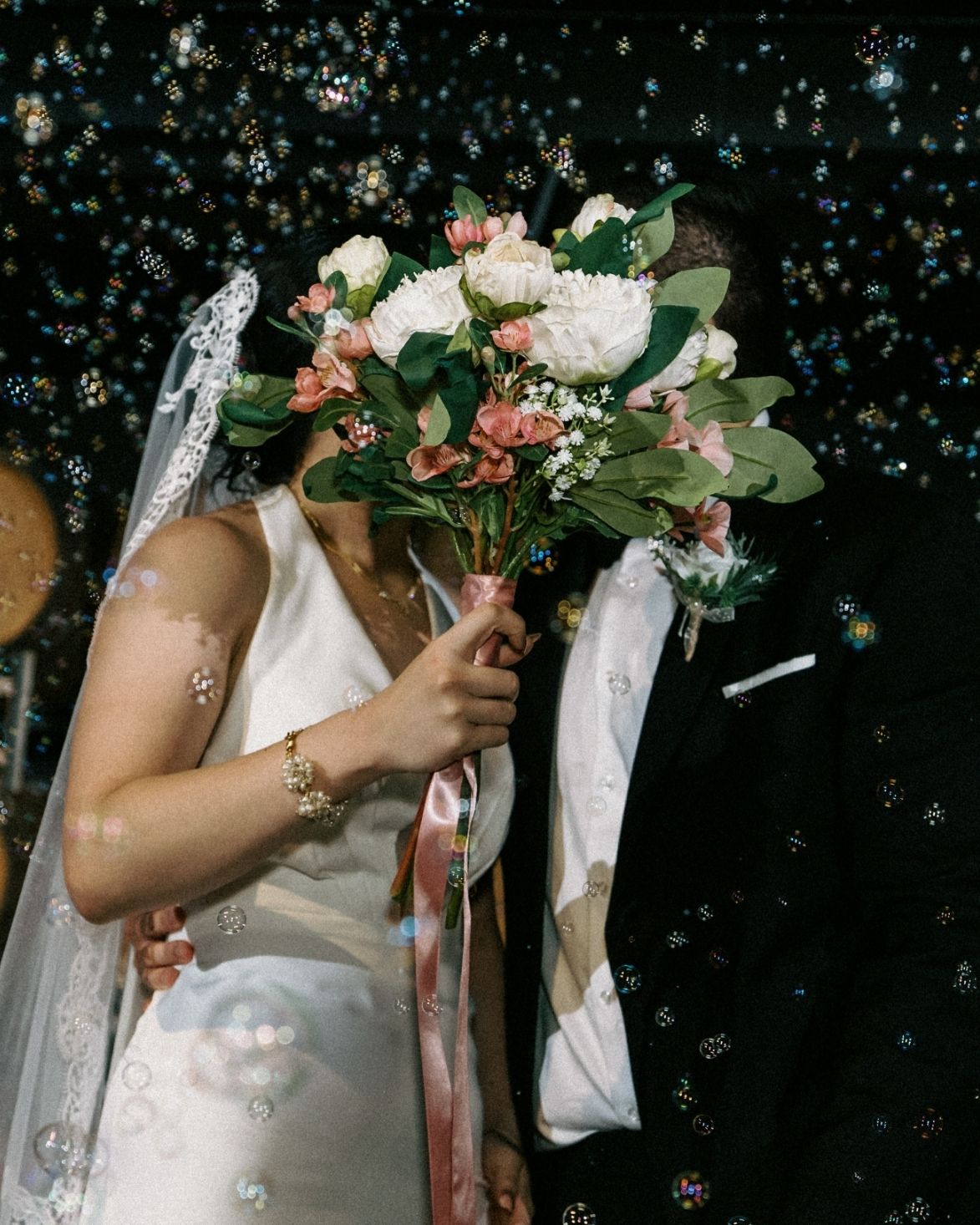Bride and groom kissing behind a white and pink wedding bouquet surrounded by floating bubbles at their reception.