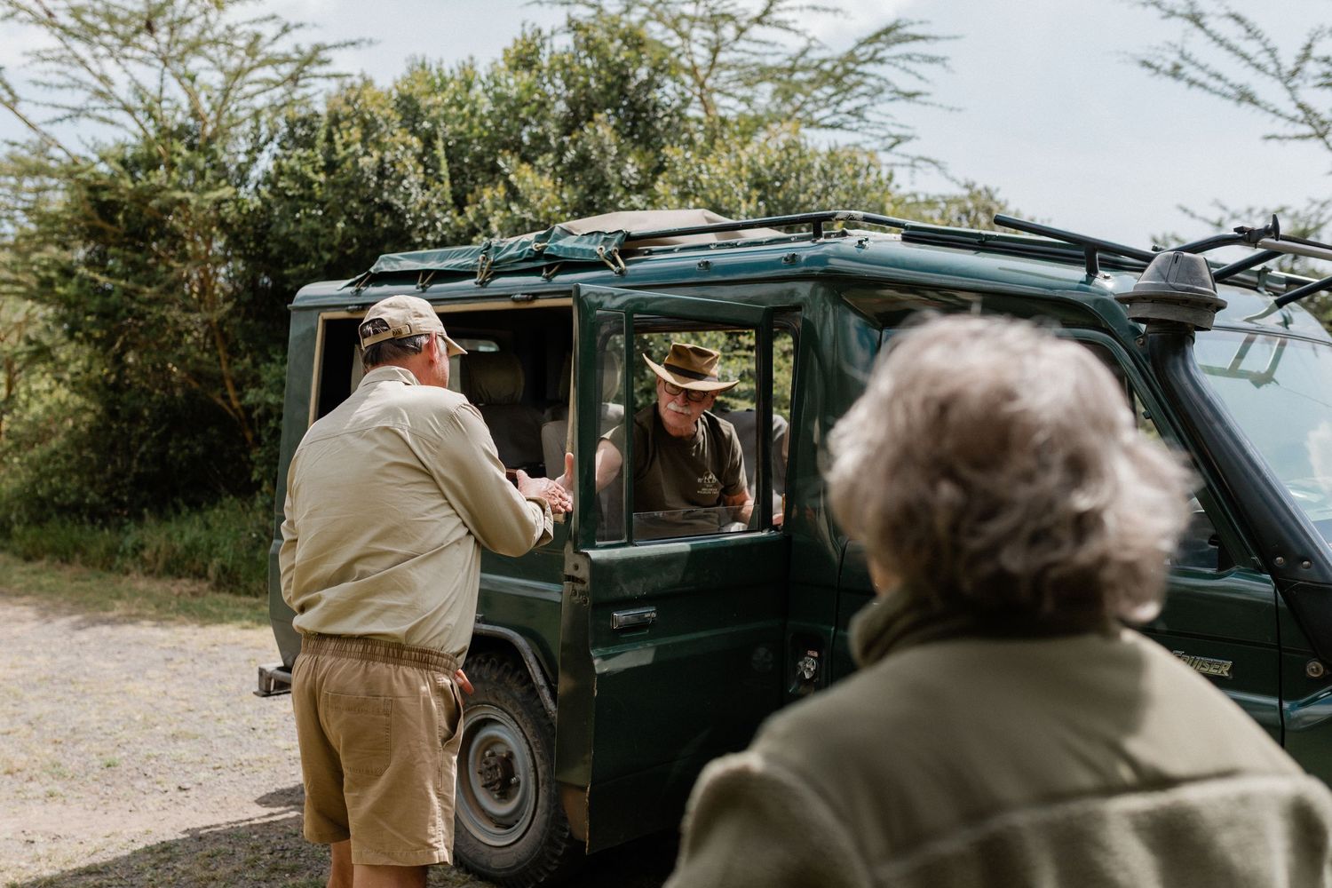 Two tourists interact with a safari guide in a green Land Rover on a dirt road surrounded by lush African vegetation.