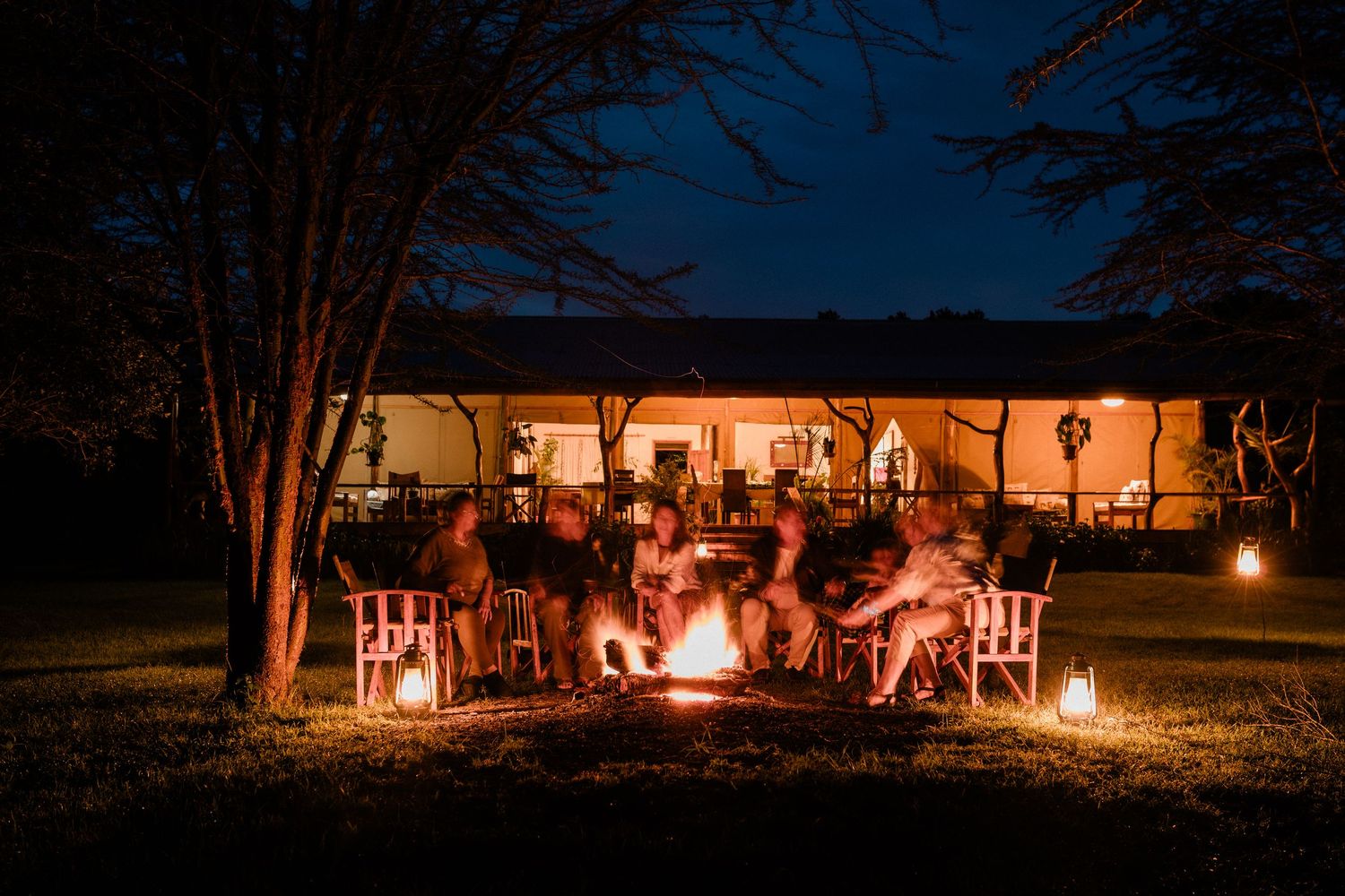 Group of people gathered around a glowing campfire at night outside a rustic lodge surrounded by trees.