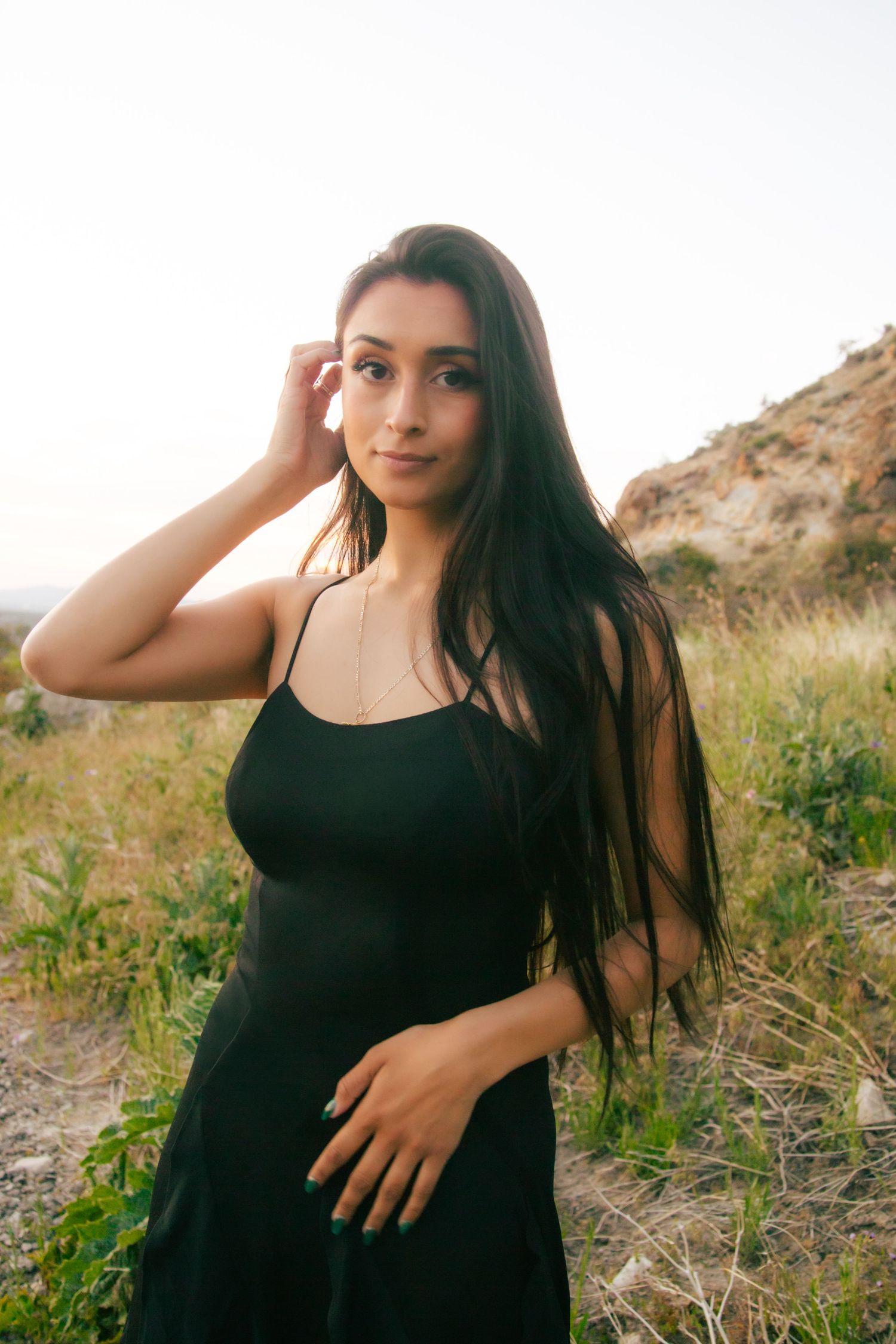 Young woman with long dark hair in black dress posing in sunlit outdoor field near hills.