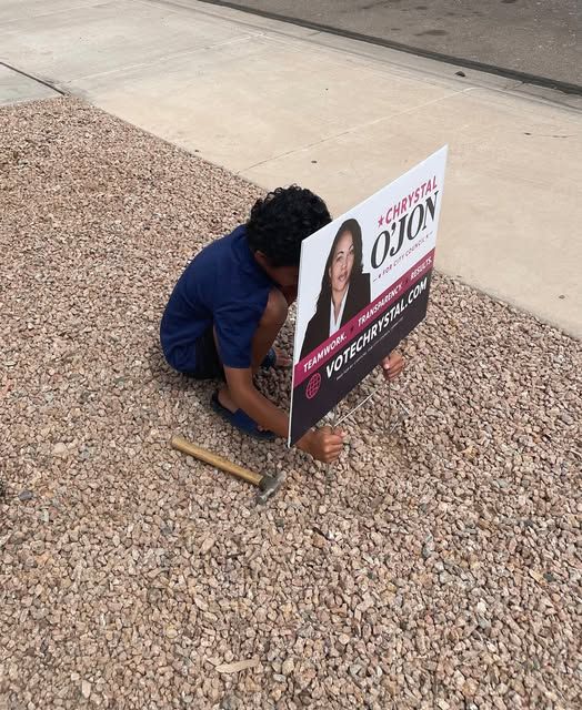 Person installing a Chrystal Ojon political campaign yard sign on gravel ground near a building.