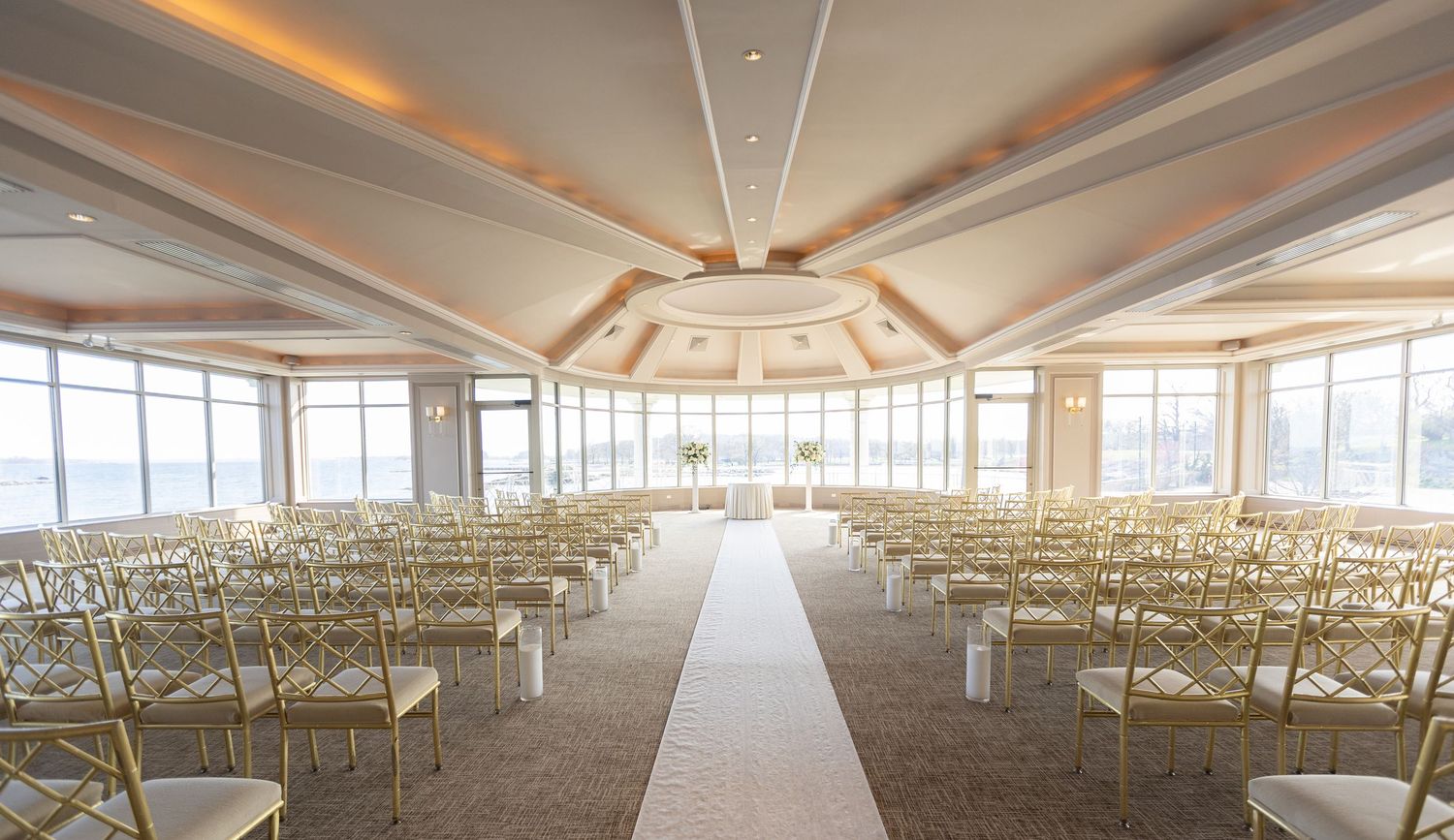 Elegant waterfront wedding ceremony setup with gold chairs and panoramic windows at a luxury wedding venue in Westchester County, New York.