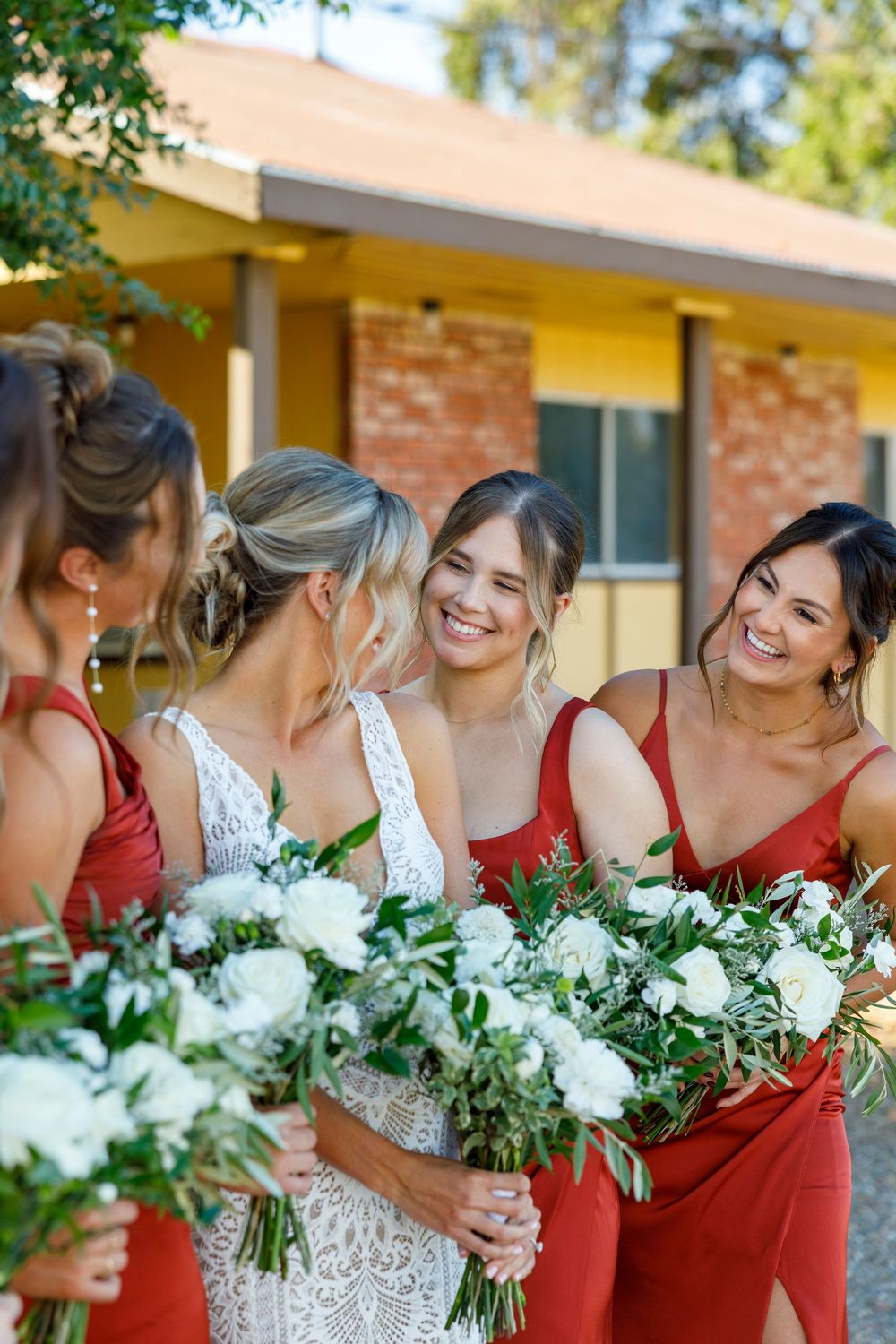 Bride in white lace gown laughing with bridesmaids in red dresses holding white floral bouquets outdoors.