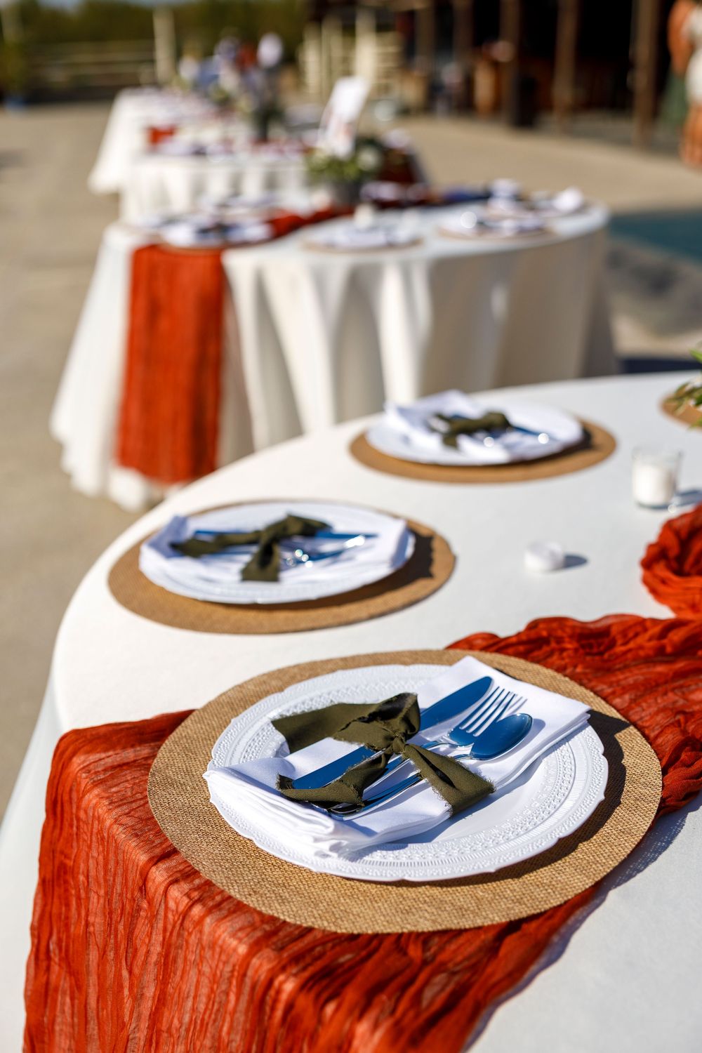 Elegant outdoor wedding table setting with rust orange runner, woven placemats, white plates, and dark green napkins.