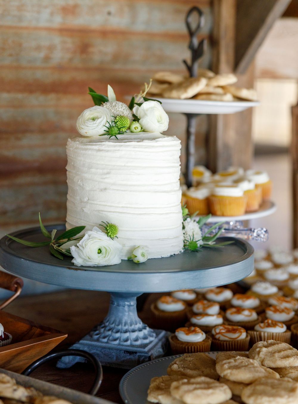 White single-tier wedding cake with white flowers on a blue pedestal stand surrounded by cookies and cupcakes.