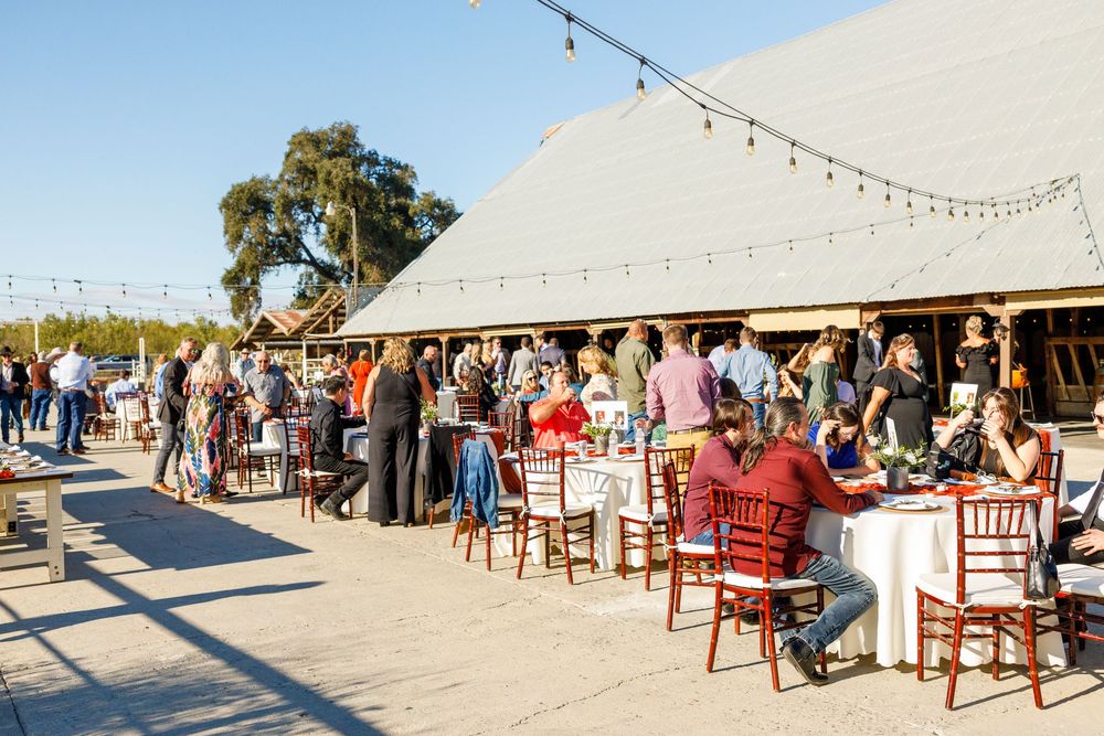 Outdoor event at a rustic barn venue with guests mingling and dining at tables under string lights.