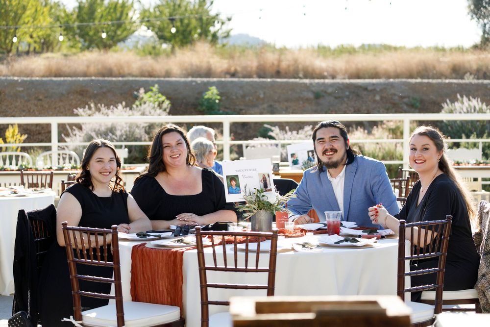 Four guests smile at an outdoor dining table set for a formal event, with scenic views in the background.