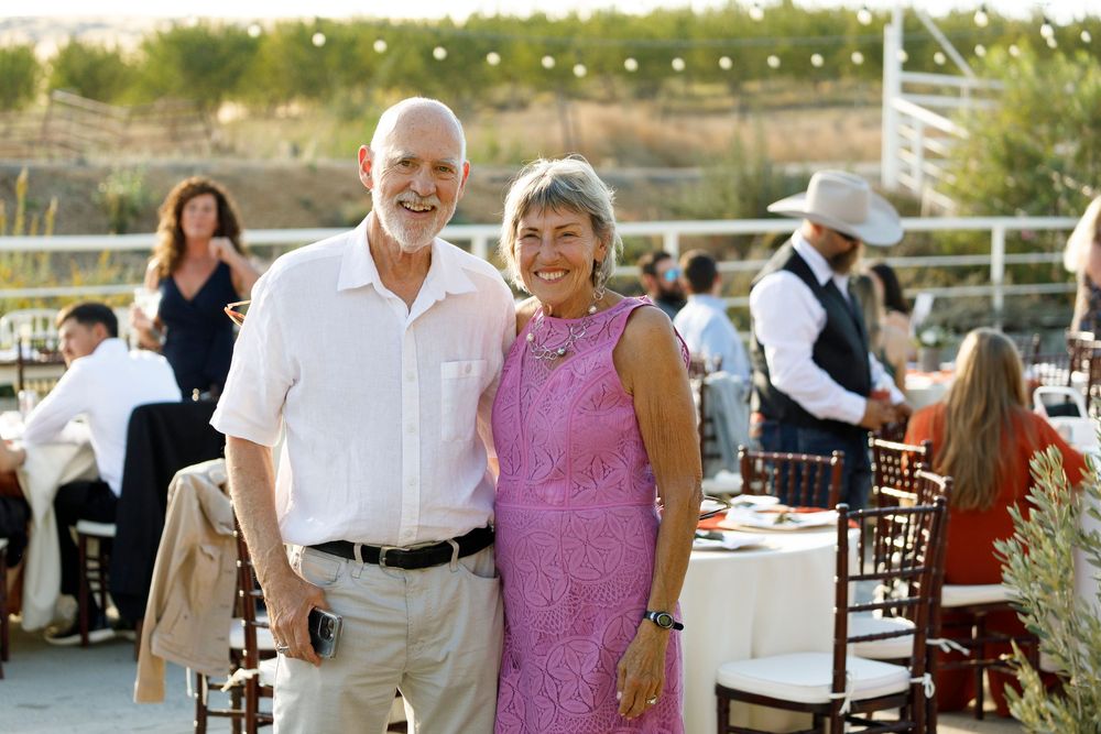 Smiling couple posing outdoors at a wedding reception with guests dining at tables in the background.