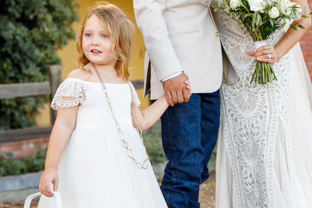 Young flower girl in white dress with pearl necklace stands beside bride and groom at outdoor wedding.