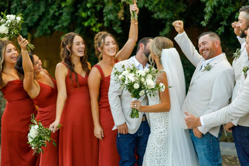 Wedding party celebrating as bride and groom kiss, bridesmaids in red dresses holding white bouquets outdoors.