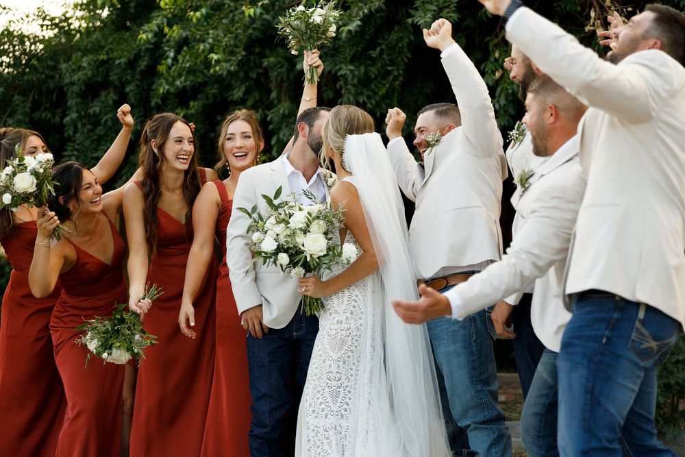 Wedding party celebrating with bride in white gown holding bouquet, bridesmaids in red dresses, groomsmen in white shirts.