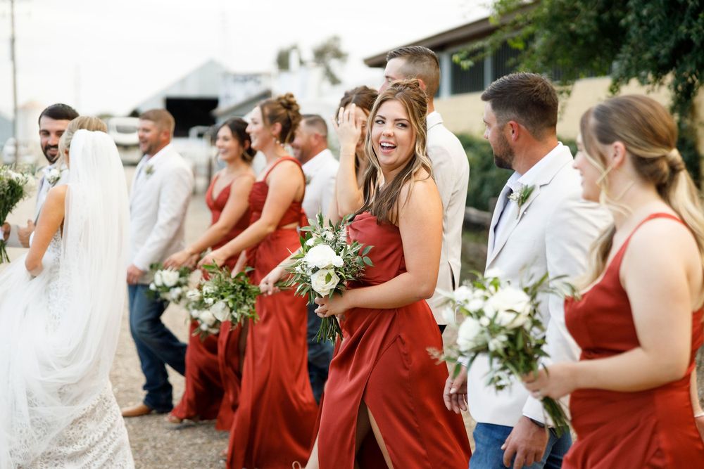 Bridesmaids in red dresses laugh joyfully while holding white floral bouquets during an outdoor wedding celebration.