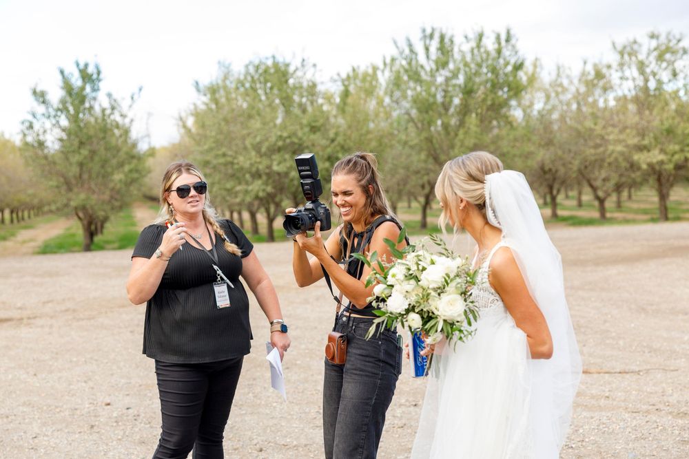 Two photographers capture a bride holding a white bouquet outdoors among trees on her wedding day.