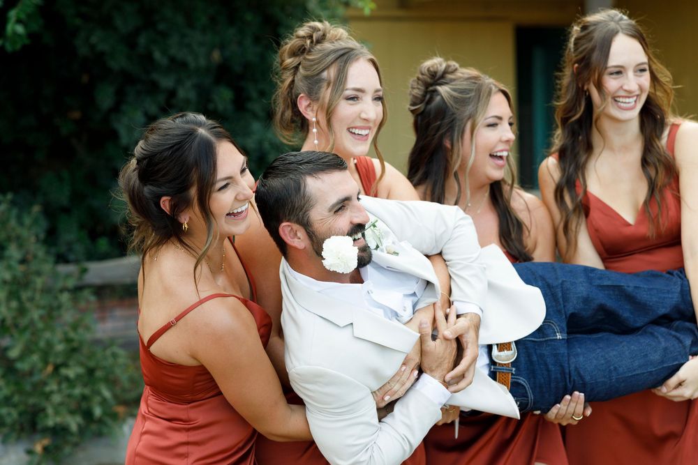 Bridesmaids in red dresses laughing while lifting groom in white suit at outdoor wedding celebration.