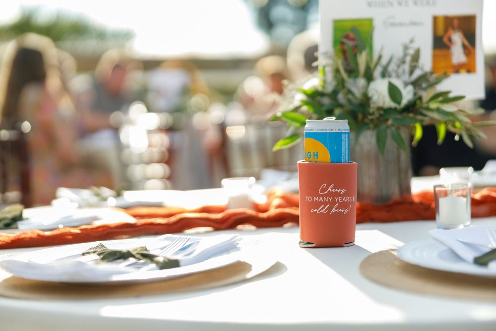 Orange koozie on a wedding reception table with floral centerpiece and place settings outdoors.