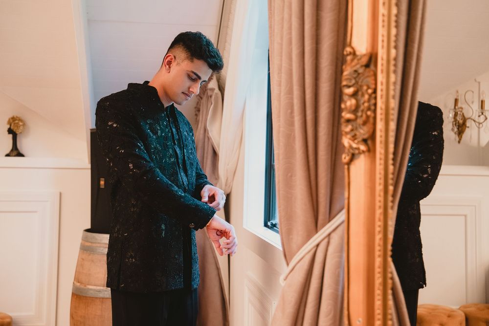 Elegant man in black embroidered jacket checking his watch by ornate gold-framed curtains indoors.