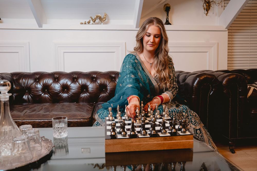 Elegant blonde woman in teal dress playing chess on glass coffee table in luxury living room.