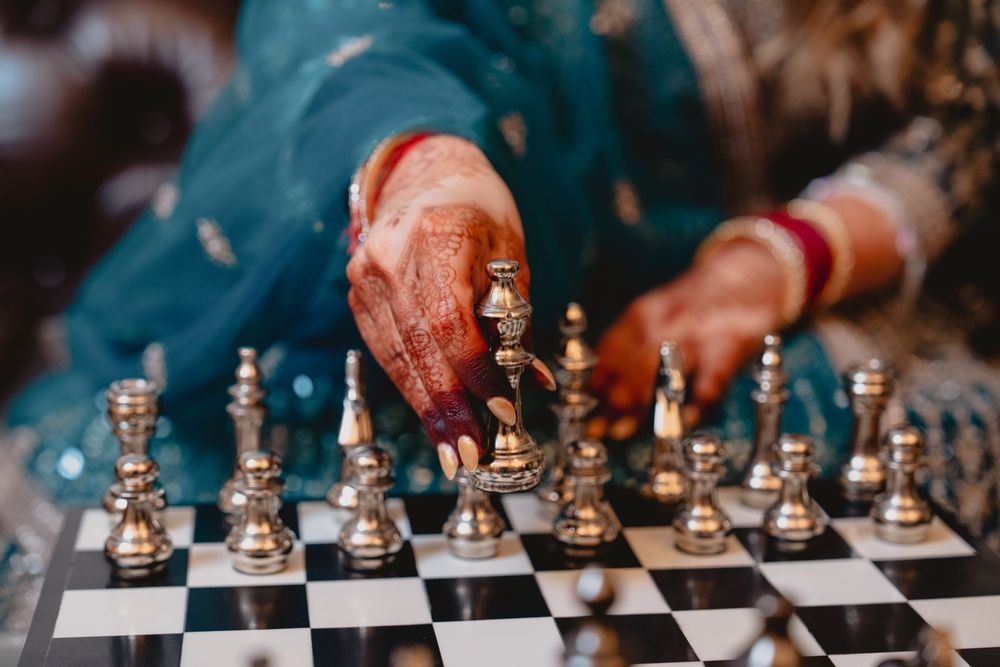A woman with henna-decorated hands moves a chess piece on a silver chess set, wearing a teal Indian outfit.