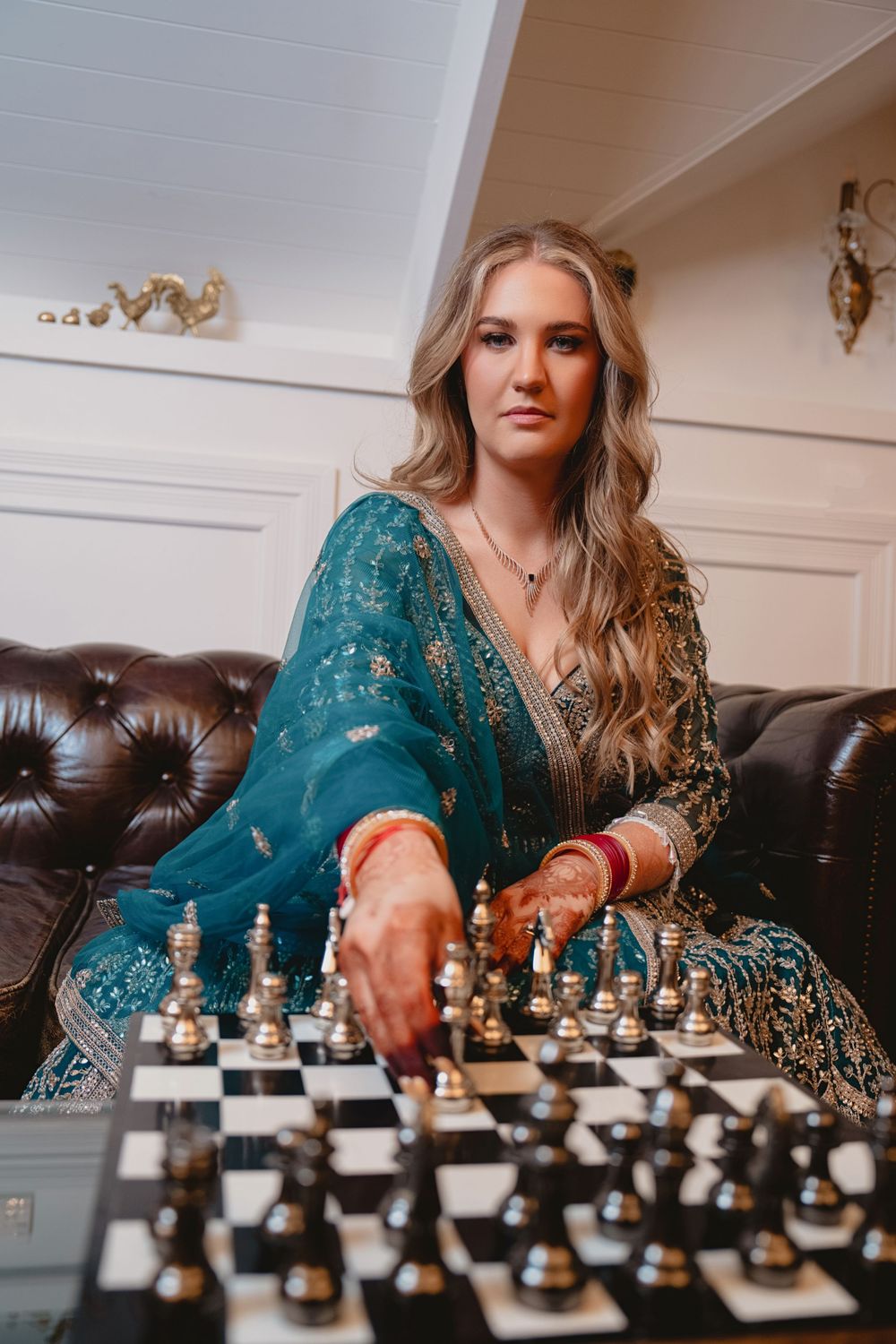 Woman in teal embroidered Indian dress with henna hands playing chess on leather sofa.