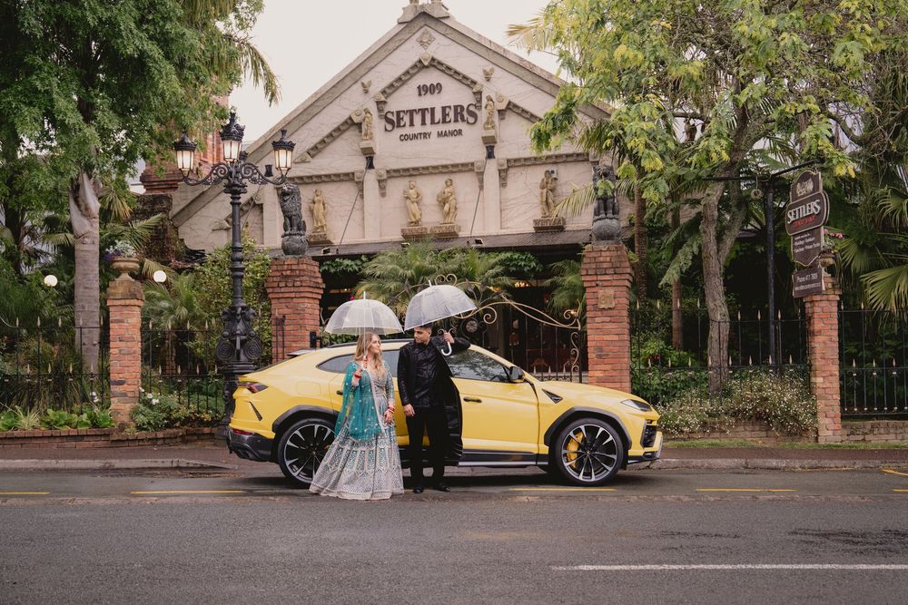 Couple with umbrellas posing beside a yellow Lamborghini Urus SUV at The Settlers estate gate entrance.