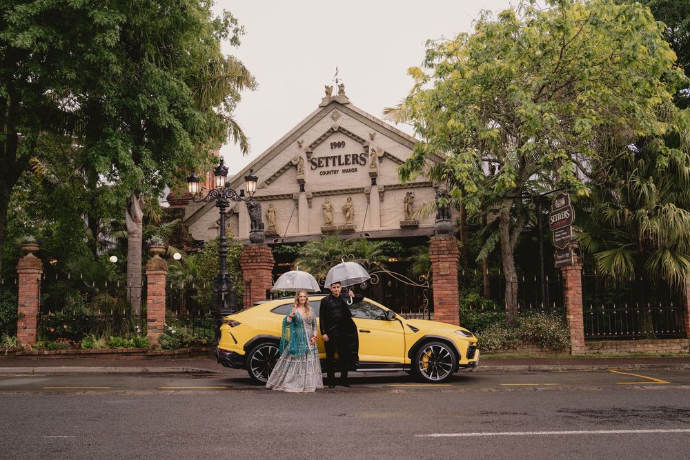 Bride and groom with white umbrellas pose before a yellow Lamborghini at Settlers venue entrance.