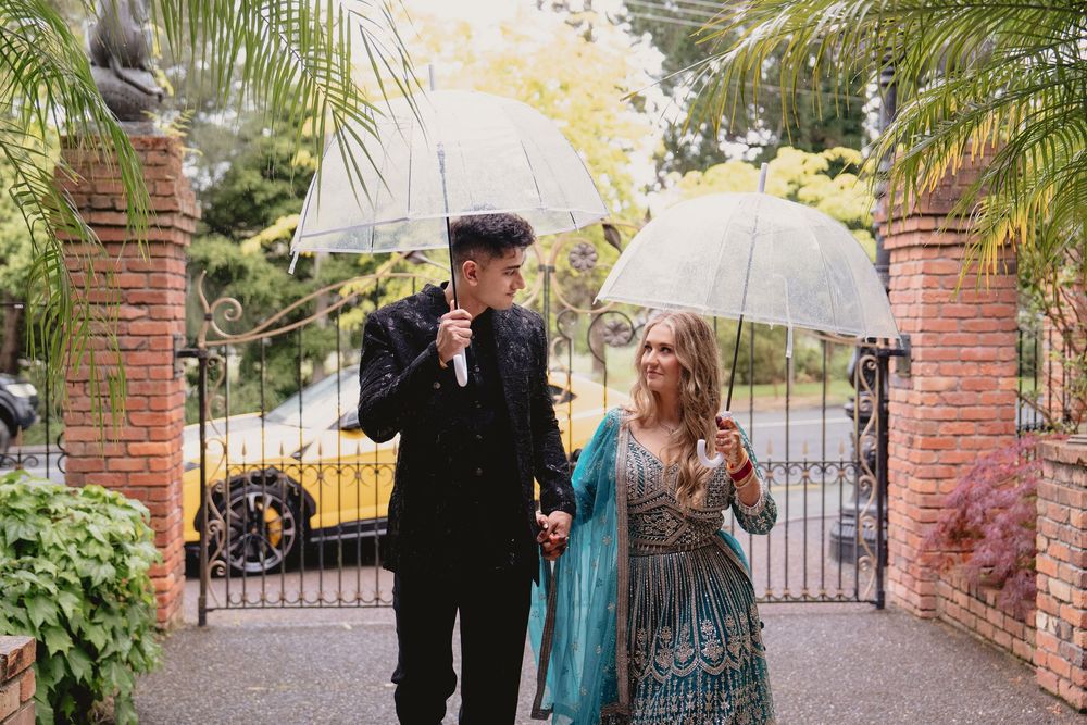 Couple walking under clear umbrellas in the rain at an elegant garden gate venue.